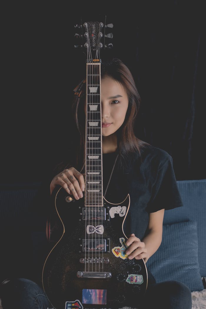 About Asian woman sitting with an electric guitar, half-covered face, in an indoor setting.